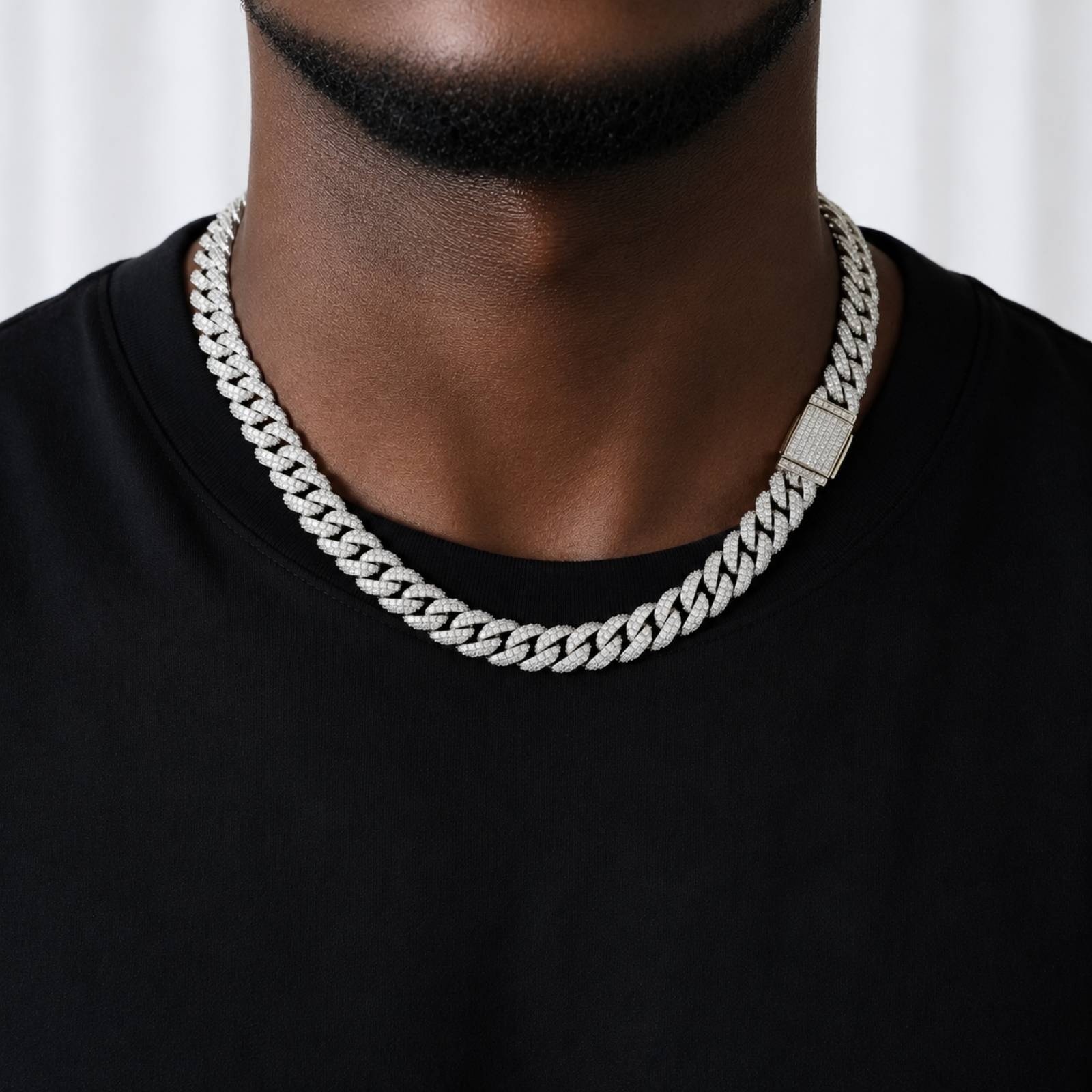 A close-up of a man wearing a thick silver Cuban link chain necklace with a rectangular pavé clasp, paired with a black t-shirt.
