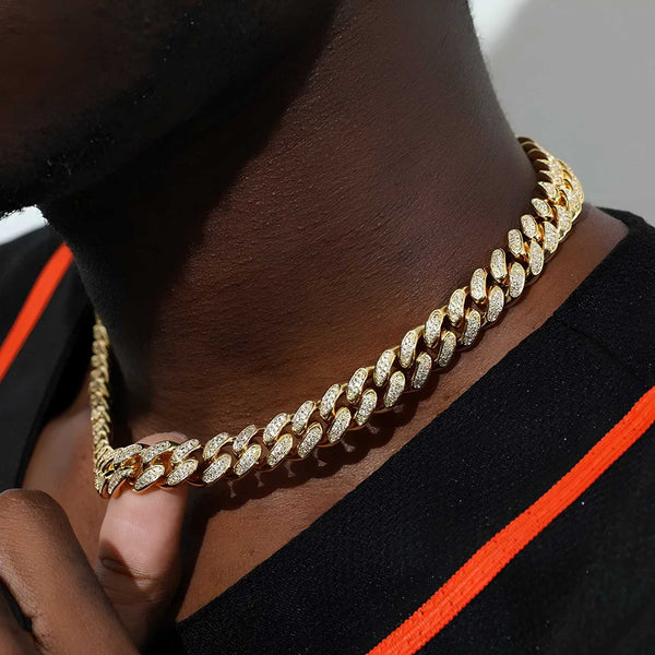 A close-up of a gold-toned Cuban link chain necklace with pavé-set clear stones worn around a person's neck, paired with a black shirt featuring a bright orange stripe.