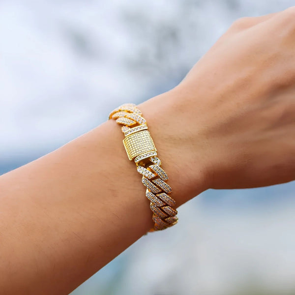 A close-up of a gold-toned cuban bracelet with a rectangular pavé clasp, worn on a person's wrist against a blurred outdoor background.