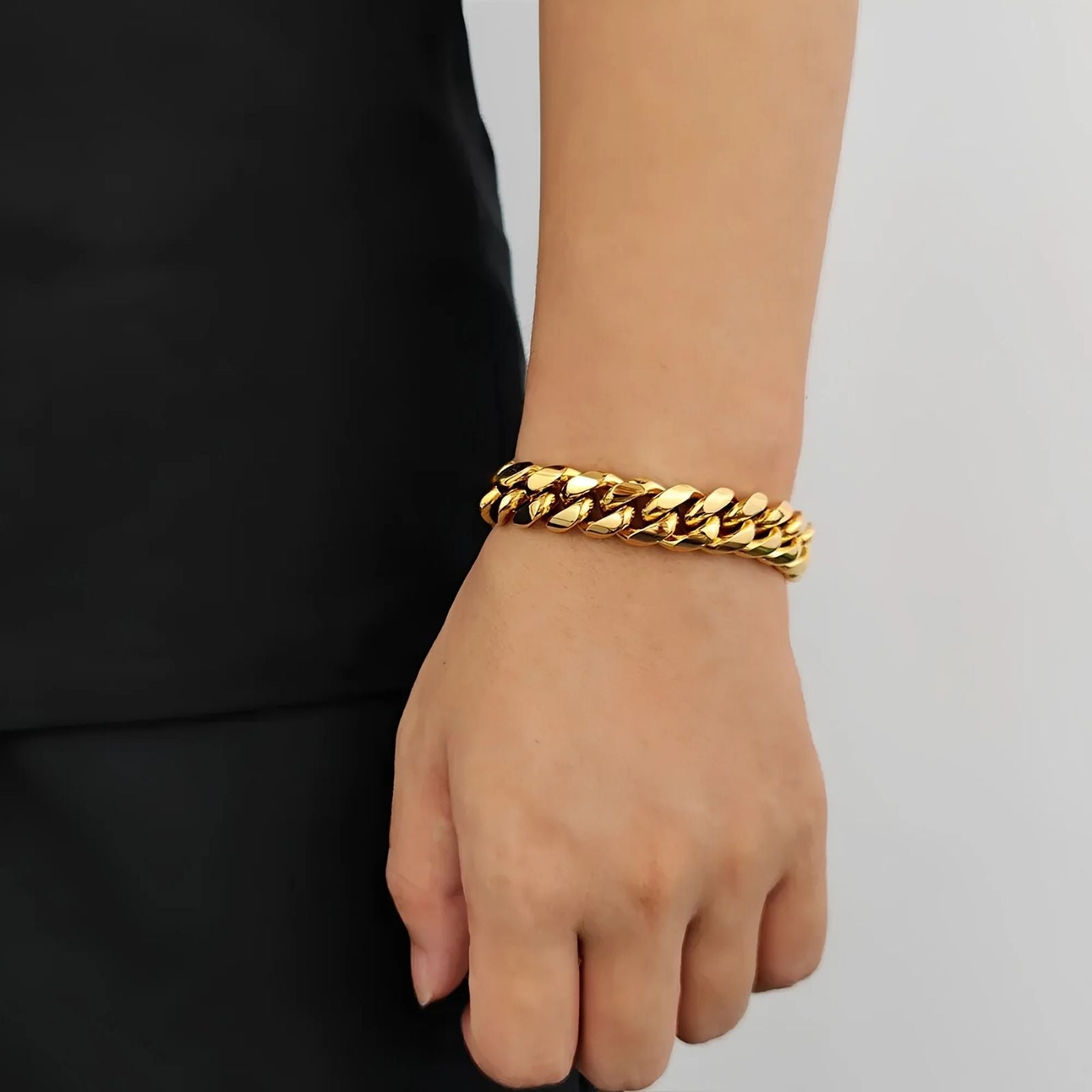A close-up of a person's wrist wearing a thick, shiny gold curb-link chain bracelet, paired with a black top against a light gray background.