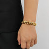 A close-up of a person's wrist wearing a thick, shiny gold curb-link chain bracelet, paired with a black top against a light gray background.
