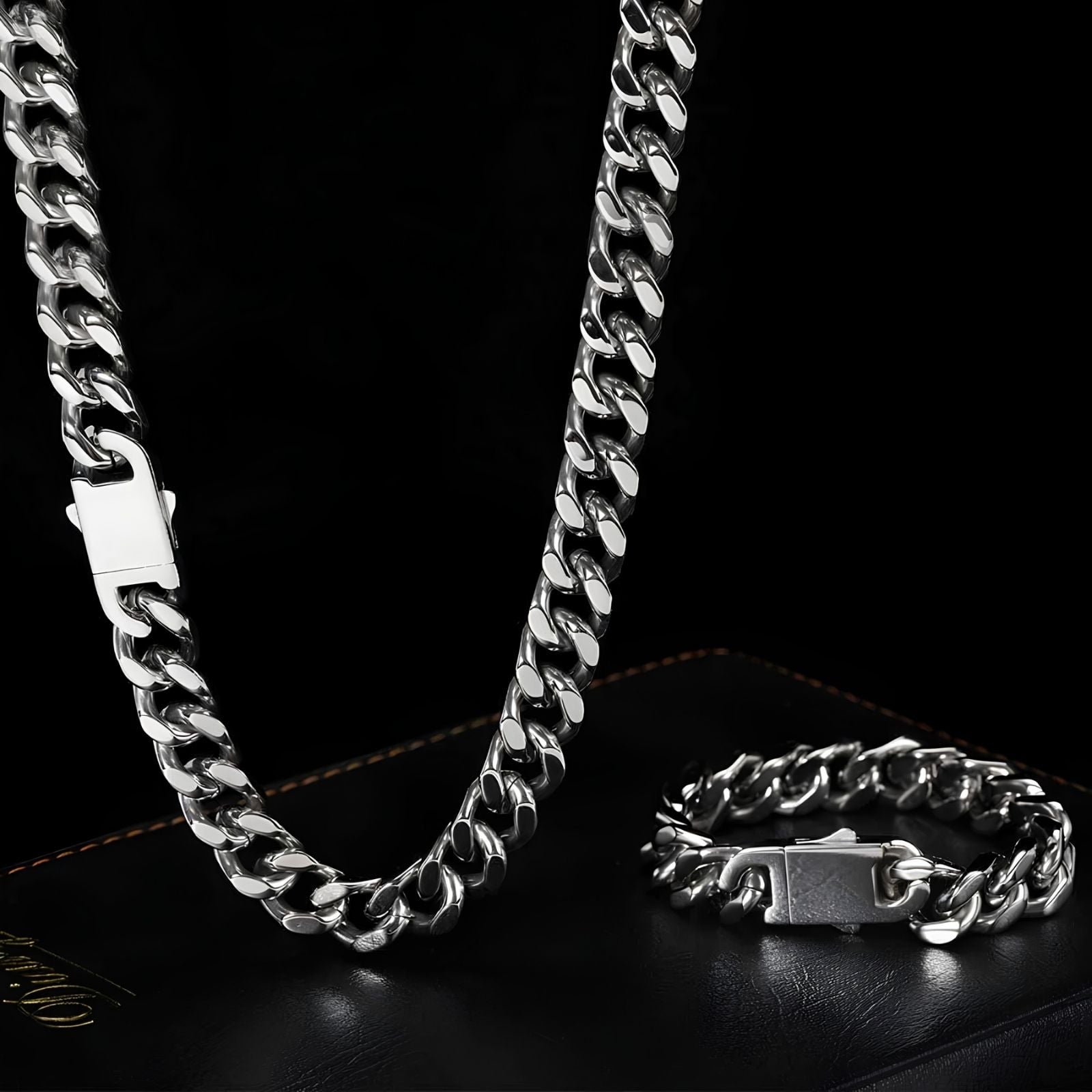 A silver-toned Cuban link chain necklace and matching bracelet displayed on a black leather surface against a black background.
