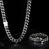 A silver-toned Cuban link chain necklace and matching bracelet displayed on a black leather surface against a black background.