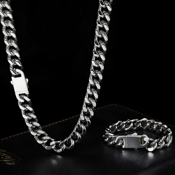 A silver-toned Cuban link chain necklace and matching bracelet displayed on a black leather surface against a black background.