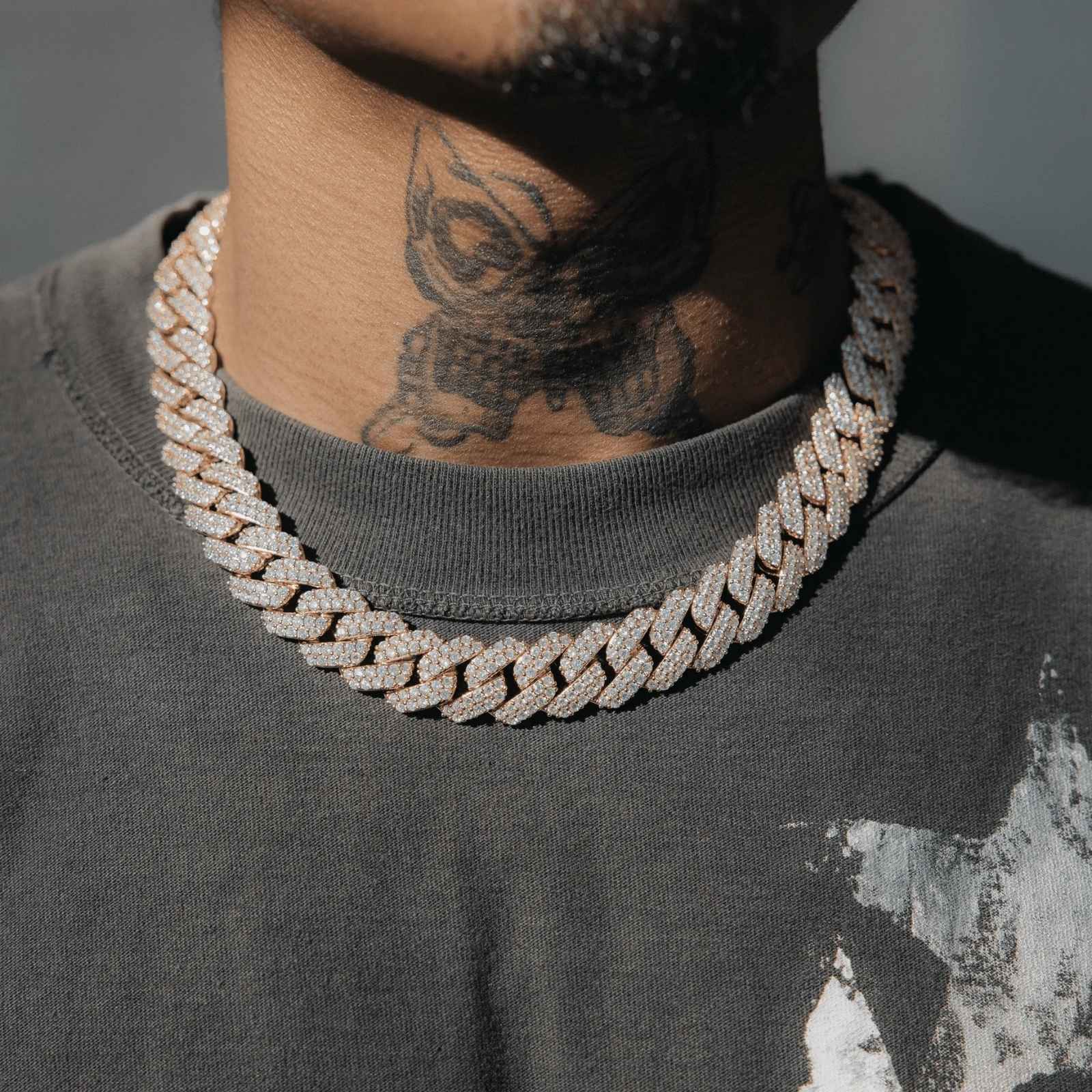 A close-up of a person wearing a thick, chunky silver-toned Cuban link chain necklace covered in small clear stones, paired with a dark gray t-shirt.