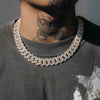 A close-up of a person wearing a thick, chunky silver-toned Cuban link chain necklace covered in small clear stones, paired with a dark gray t-shirt.
