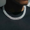 A close-up of a man wearing a thick, silver-toned Cuban link chain necklace paved with small clear stones, styled over a black t-shirt.