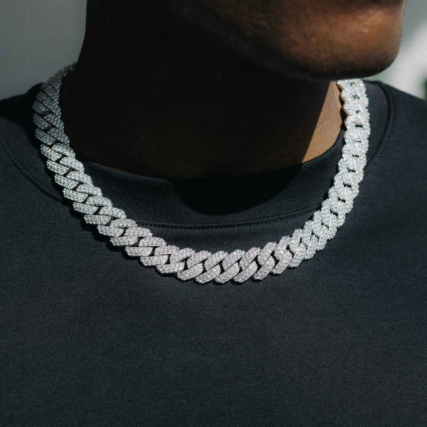 A close-up of a man wearing a thick, silver-toned Cuban link chain necklace paved with small clear stones, styled over a black t-shirt.