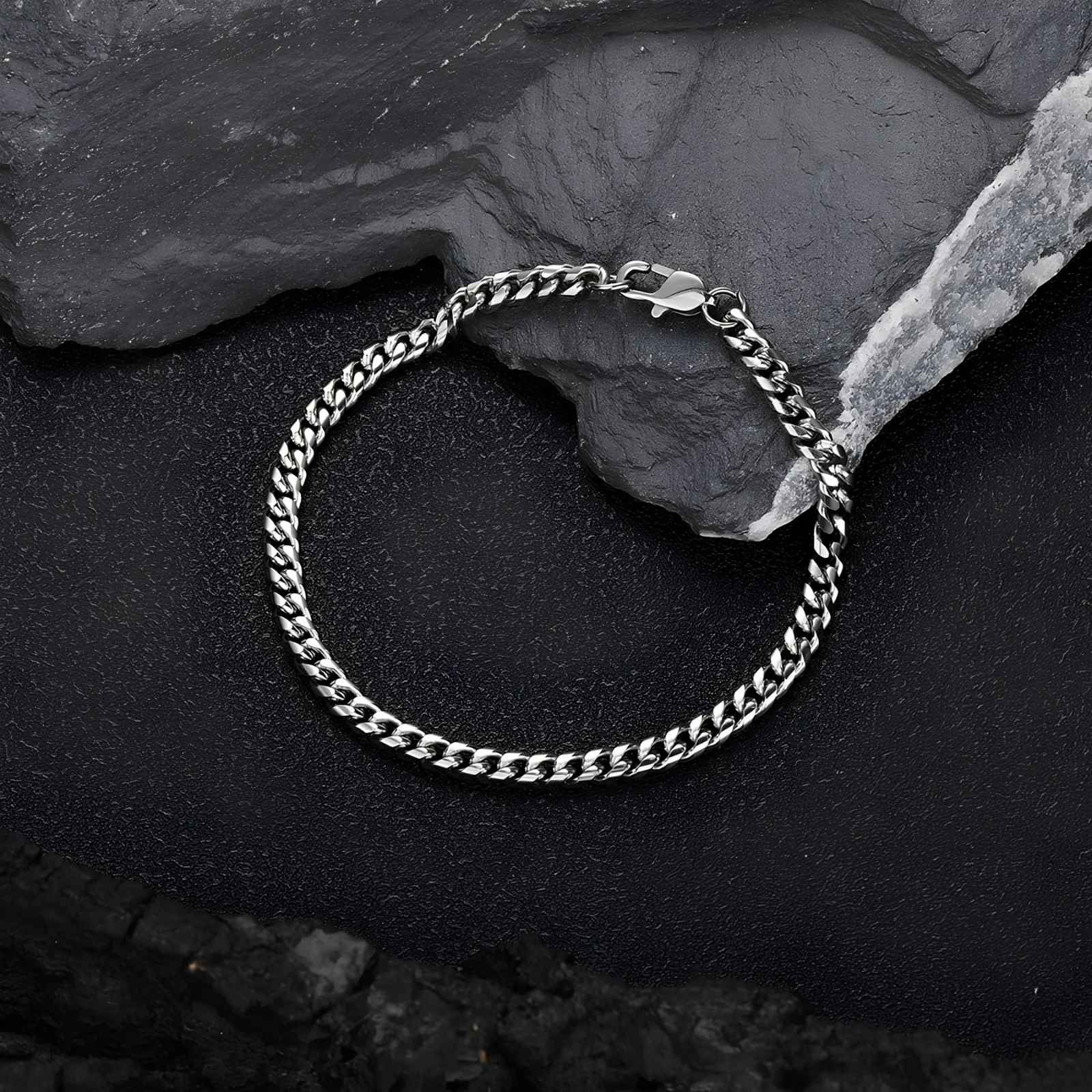 A silver-toned metal chain bracelet with a curb link design and a spring ring clasp, displayed on a textured black background with dark slate stones.