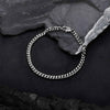 A silver-toned metal chain bracelet with a curb link design and a spring ring clasp, displayed on a textured black background with dark slate stones.