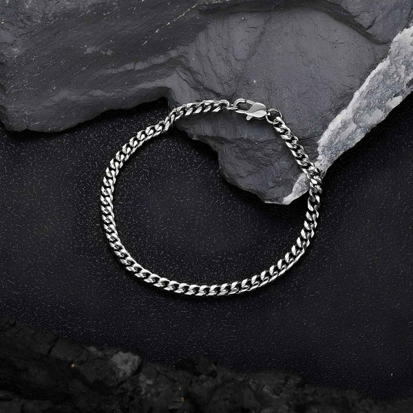 A silver-toned metal chain bracelet with a curb link design and a spring ring clasp, displayed on a textured black background with dark slate stones.
