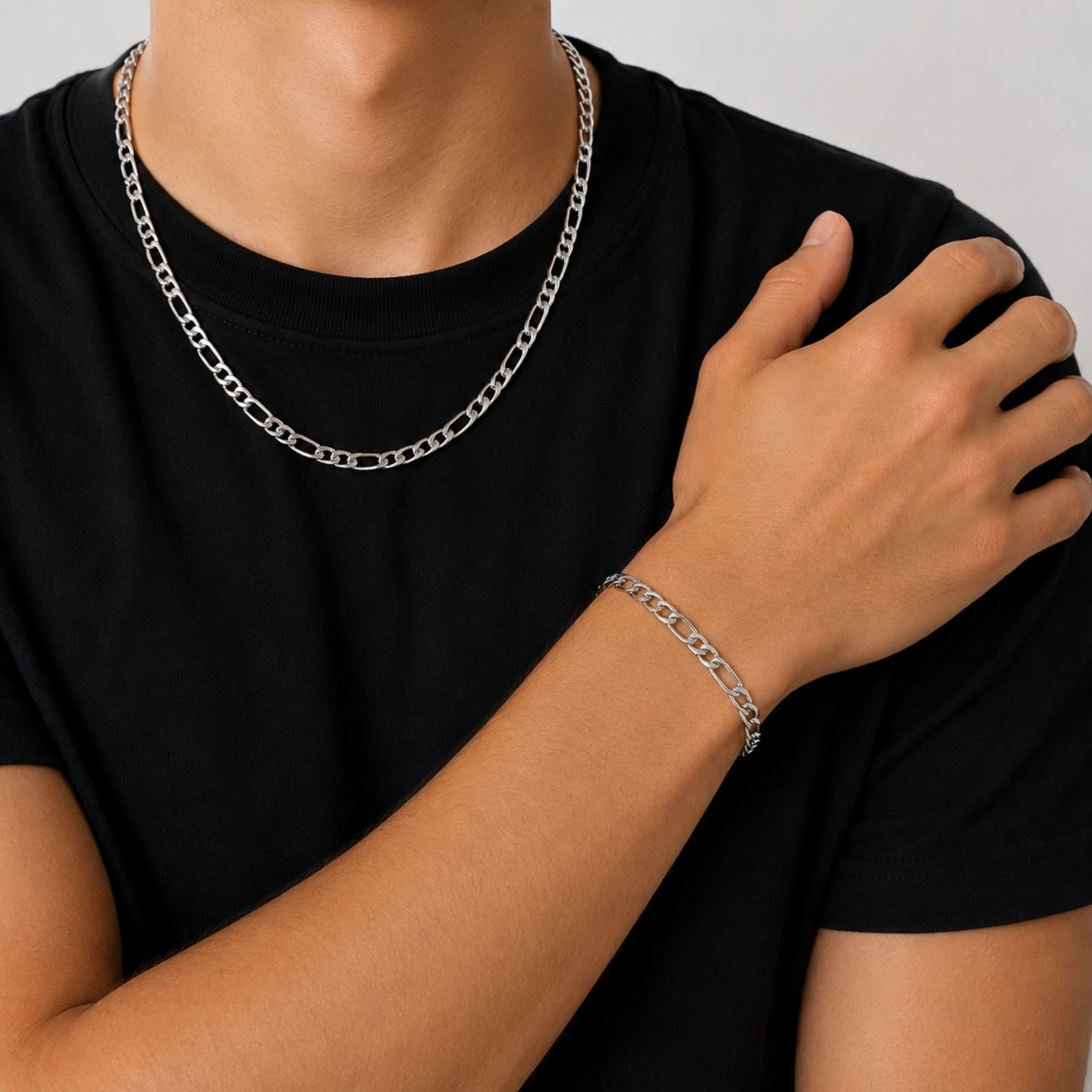 A close-up of a person wearing a silver chain necklace and a matching silver chain bracelet, paired with a black t-shirt against a light gray background.