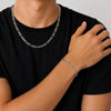 A close-up of a person wearing a silver chain necklace and a matching silver chain bracelet, paired with a black t-shirt against a light gray background.
