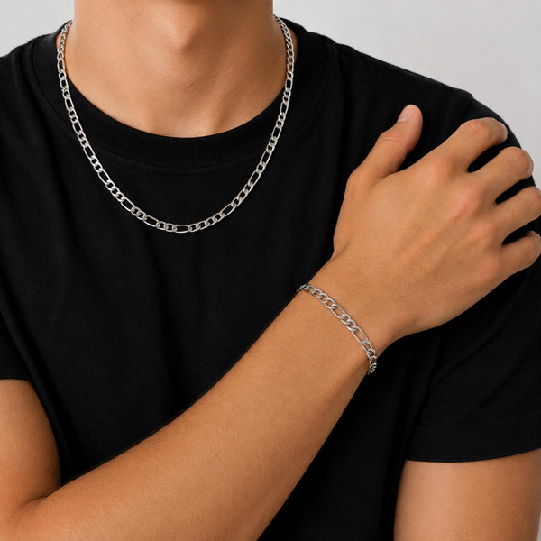 A close-up of a person wearing a silver chain necklace and a matching silver chain bracelet, paired with a black t-shirt against a light gray background.