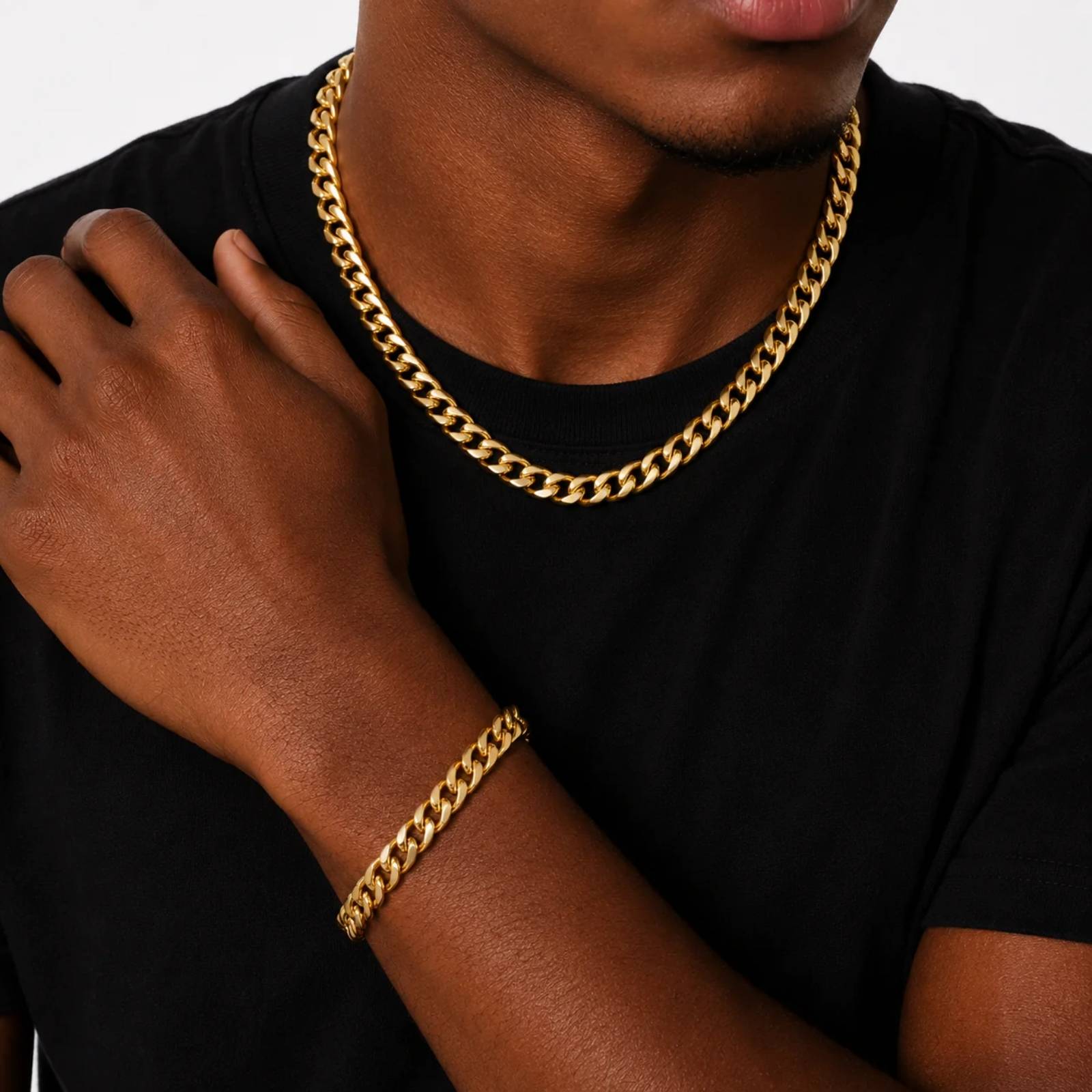 A close-up of a person wearing a thick gold Cuban link chain necklace and a matching gold bracelet, paired with a black t-shirt against a light gray background.