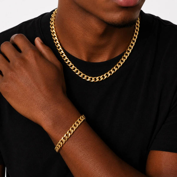 A close-up of a person wearing a thick gold Cuban link chain necklace and a matching gold bracelet, paired with a black t-shirt against a light gray background.