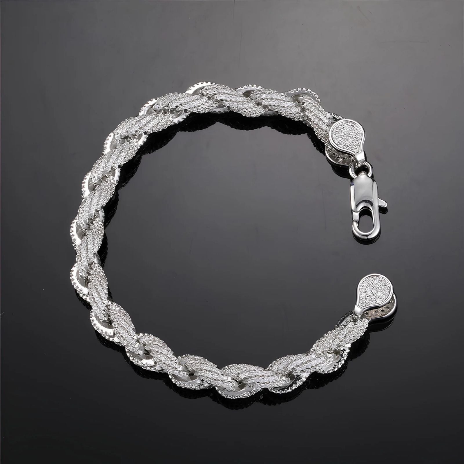 A silver-toned braided chain bracelet with pavé-set clear stones, featuring a lobster clasp and matching round end caps, displayed on a dark reflective surface.