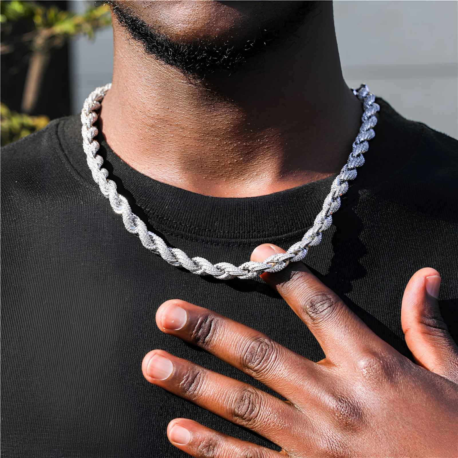 A close-up of a man wearing a chunky, silver-toned rope style chain necklace over a black t-shirt, with his hand resting on his chest.