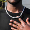 A close-up of a man wearing a chunky, silver-toned rope style chain necklace over a black t-shirt, with his hand resting on his chest.