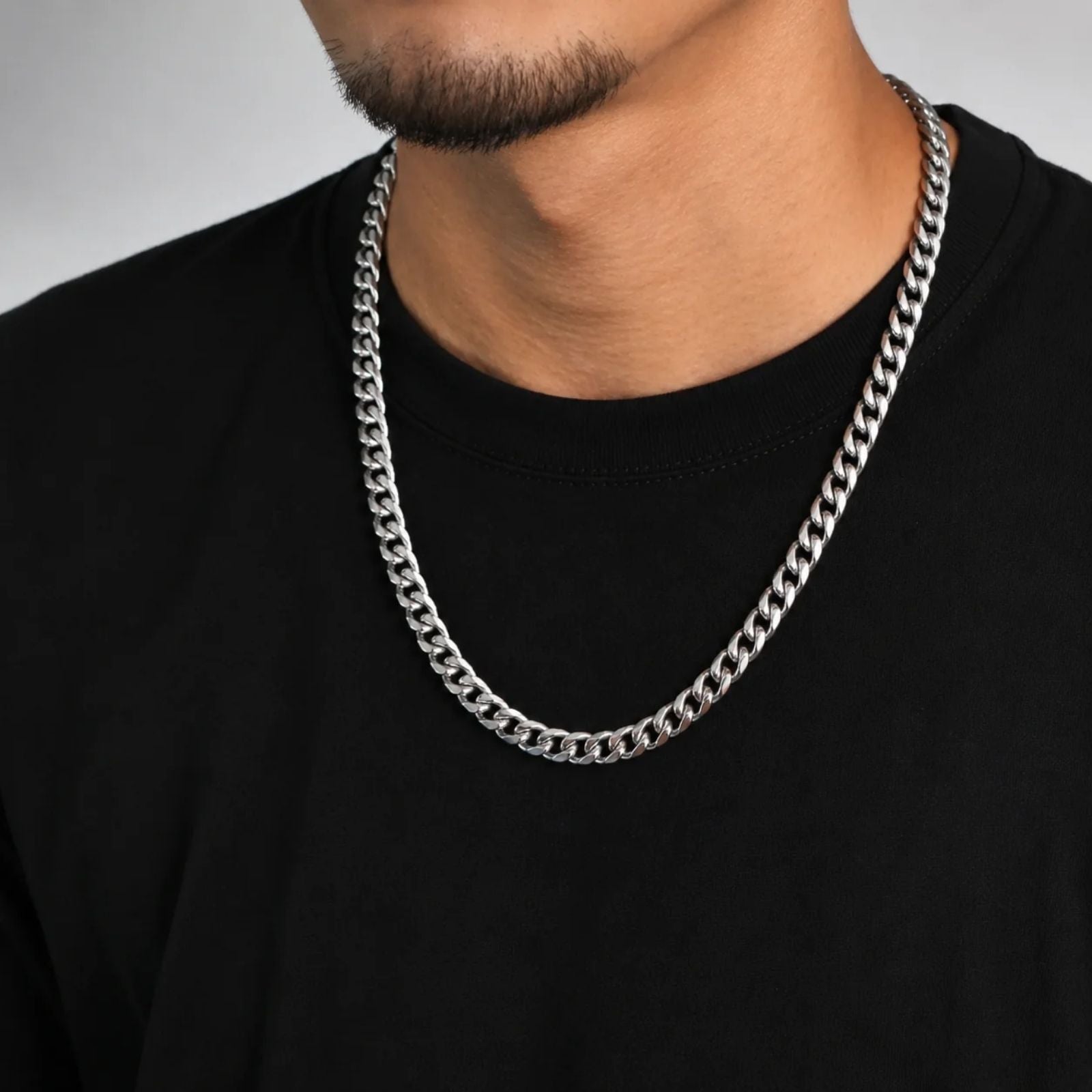 A close-up of a man wearing a thick silver Cuban link chain necklace over a black t-shirt, cropped from the chin down.