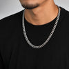 A close-up of a man wearing a thick silver Cuban link chain necklace over a black t-shirt, cropped from the chin down.