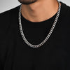 A close-up of a man wearing a thick silver Cuban link chain necklace over a black t-shirt, cropped from the chin down.