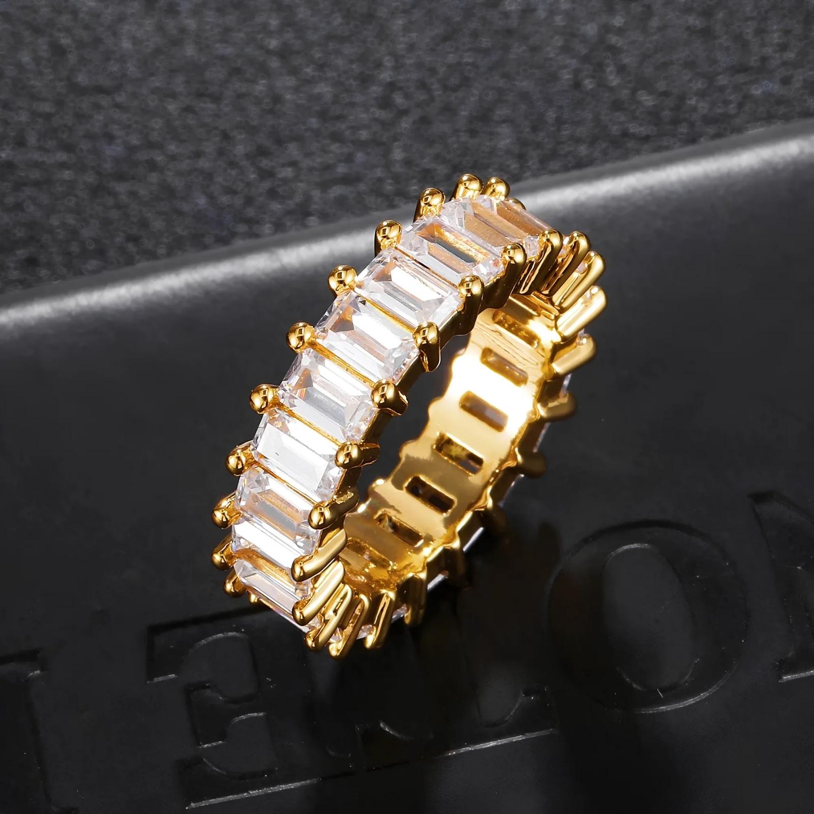 A gold-plated eternity ring featuring a row of clear rectangular baguette-cut cubic zirconia, displayed on a textured black surface.