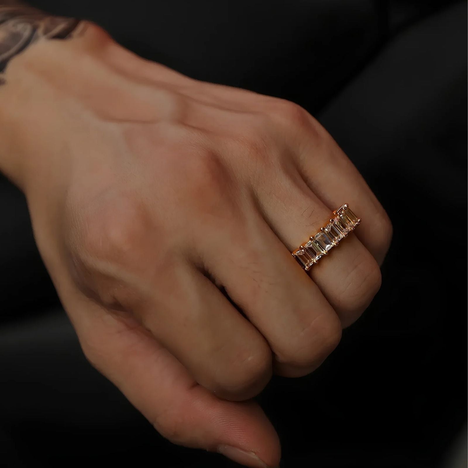 A close-up of a person's hand wearing a gold eternity ring with rectangular clear cubic zirconia, set against a dark background.