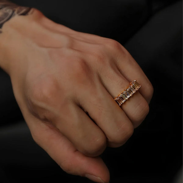 A close-up of a person's hand wearing a gold eternity ring with rectangular clear cubic zirconia, set against a dark background.