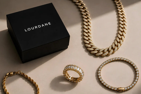 Luxury Lourdane jewellery flat lay featuring a black Lourdane jewellery box, gold iced Cuban chain, gold tennis bracelet, gold rope bracelet and iced eternity ring on a clean beige background.