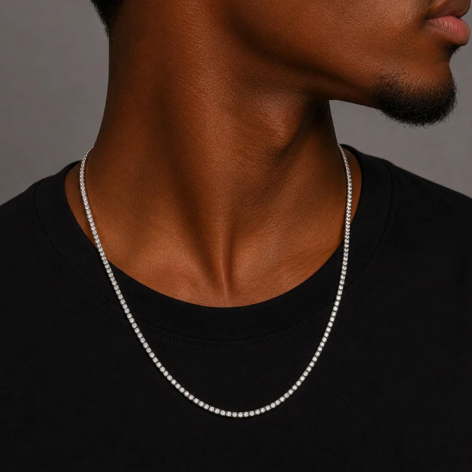 A close-up of a man wearing a delicate silver chain tennis necklace over a black t-shirt, showing the necklace from the chest up against a neutral gray background.