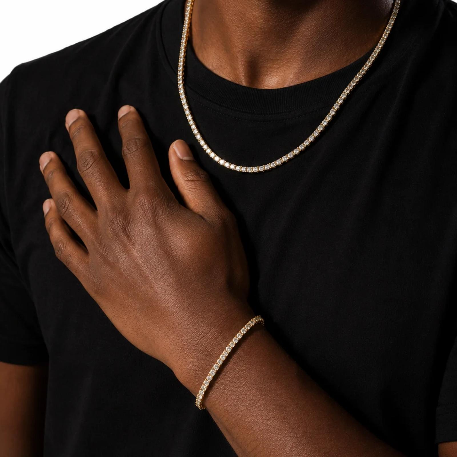 A close-up of a person wearing a gold chain necklace and a matching gold bracelet, with the hand resting on the chest over a black t-shirt.