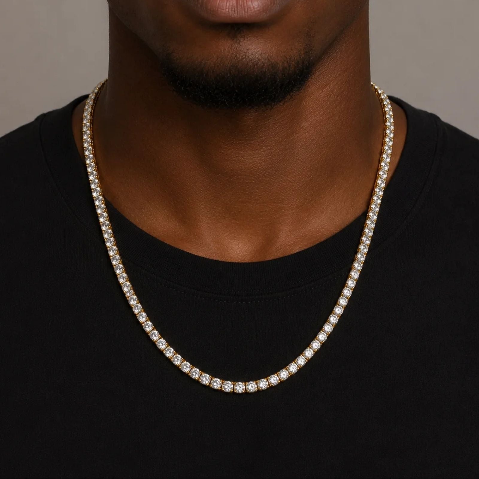 A close-up of a man wearing a gold-toned tennis chain necklace paved with clear cubic zirconia stones, styled over a black t-shirt.