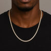 A close-up of a man wearing a gold-toned tennis chain necklace paved with clear cubic zirconia stones, styled over a black t-shirt.