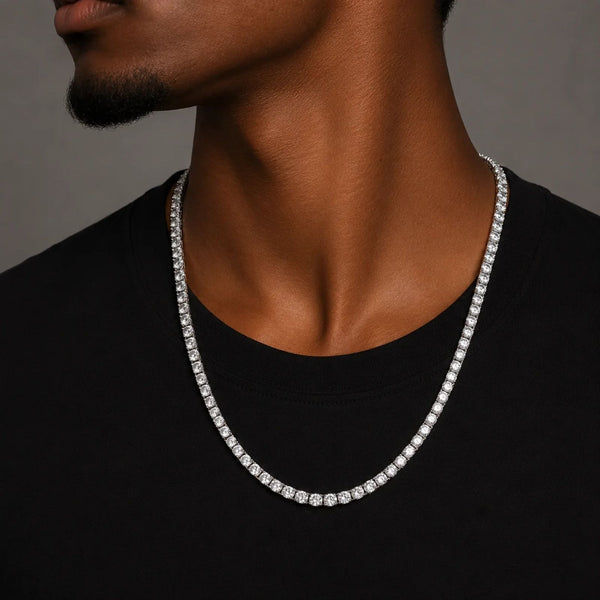 A close-up of a man wearing a silver-toned tennis chain necklace with clear stones, styled with a black t-shirt against a neutral gray background.