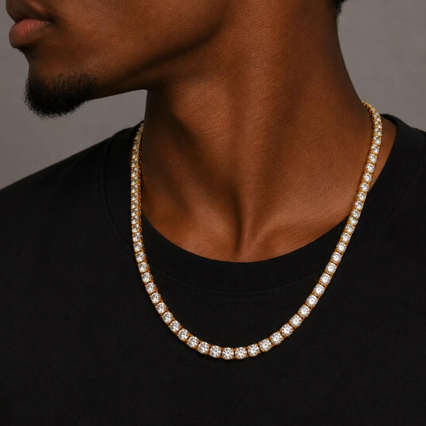 A close-up of a man wearing a gold-toned tennis necklace paved with clear cubic zirconia, styled with a black t-shirt against a neutral gray background.