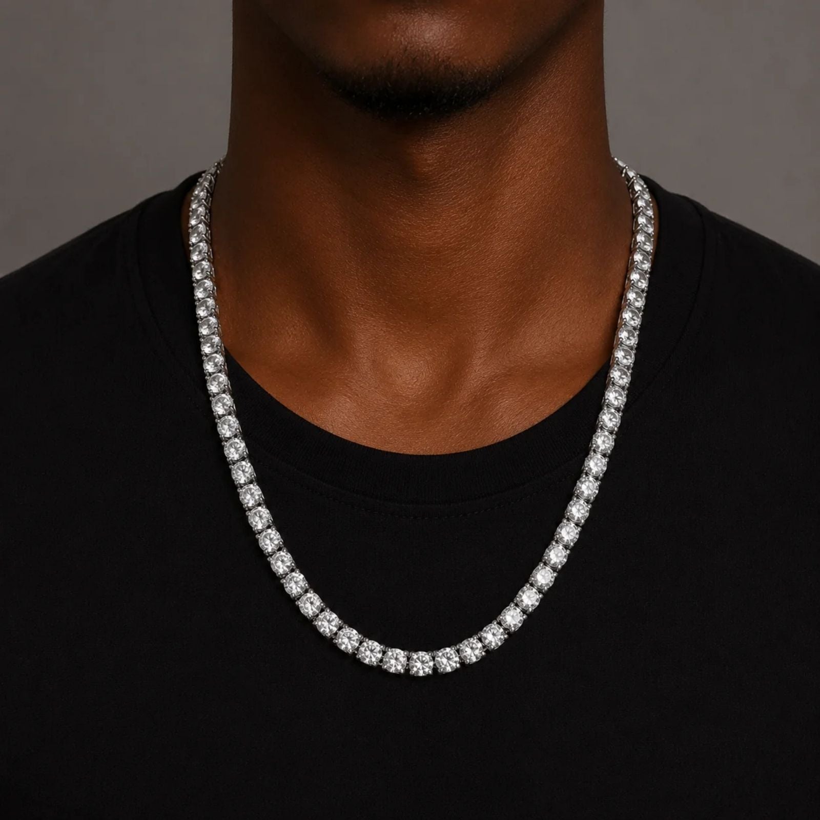 A close-up of a man wearing a silver-toned tennis chain necklace over a black t-shirt, set against a neutral gray background.
