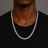 A close-up of a man wearing a silver-toned tennis chain necklace over a black t-shirt, set against a neutral gray background.