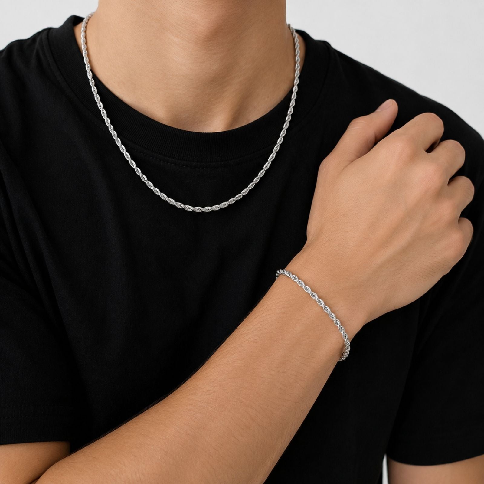 A close-up of a person wearing a silver twisted rope chain necklace and a matching silver bracelet, styled with a black t-shirt against a light gray background.